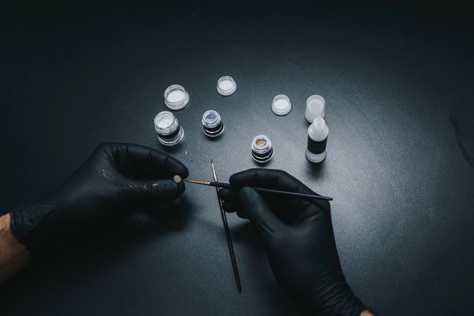 Ceramist hands with a fine brush and stain pots on a black bench in the ACE DNTL laboratory